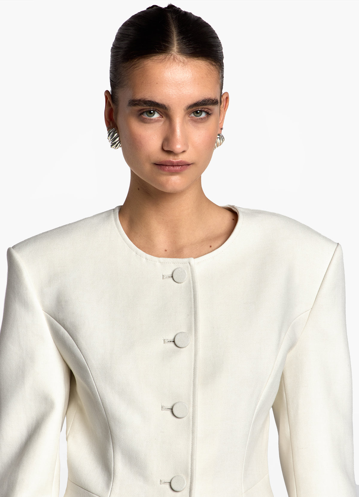 Close up shot of model wearing cream fitted wool jacket, showing round necklines and fabric covered buttons.