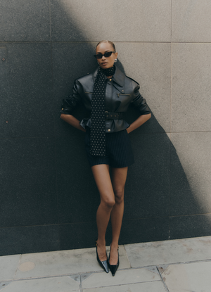 Model leaning against wall front facing. Wearing black leather cropped jacket and navy pinstripe shorts outside in the city.